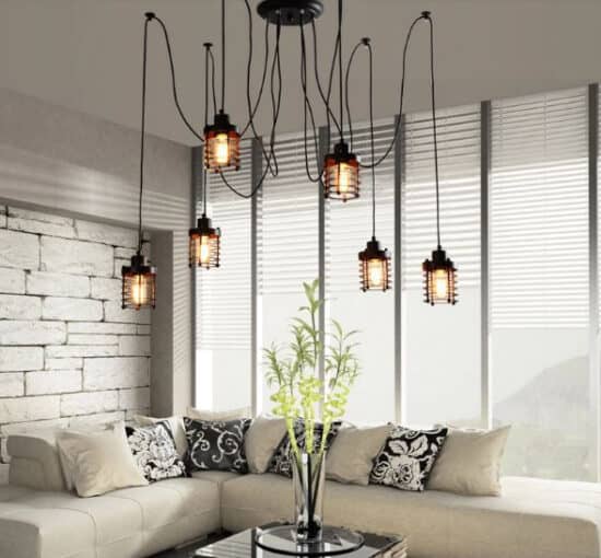 Aina Web Pendant Light - A black, industrial-style chandelier with multiple cage-like shades and Edison bulbs. The chandelier is hanging over a living room with a sofa, coffee table, and a brick wall.