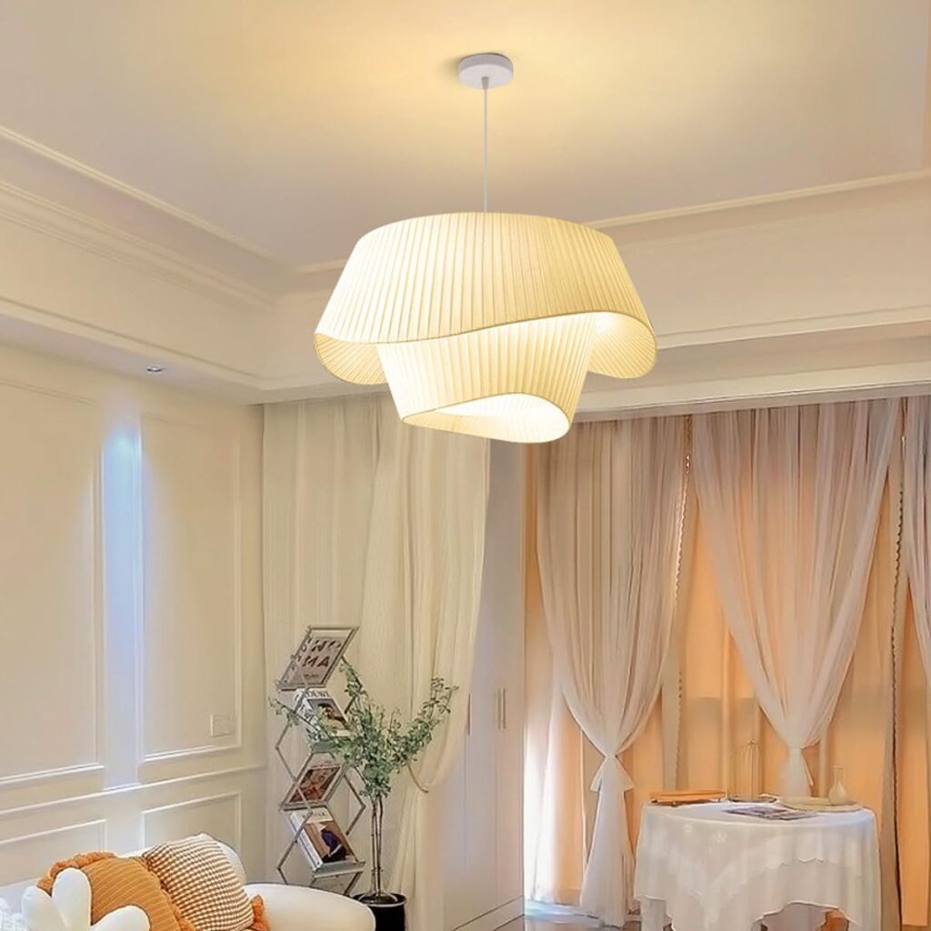 Somerled Smooth Pleated Cloth Pendant Light - A modern living room with a large, white, wave-shaped pendant light hanging from the ceiling. The light casts a warm, ambient glow onto the white sofa, potted plant, and TV in the background.