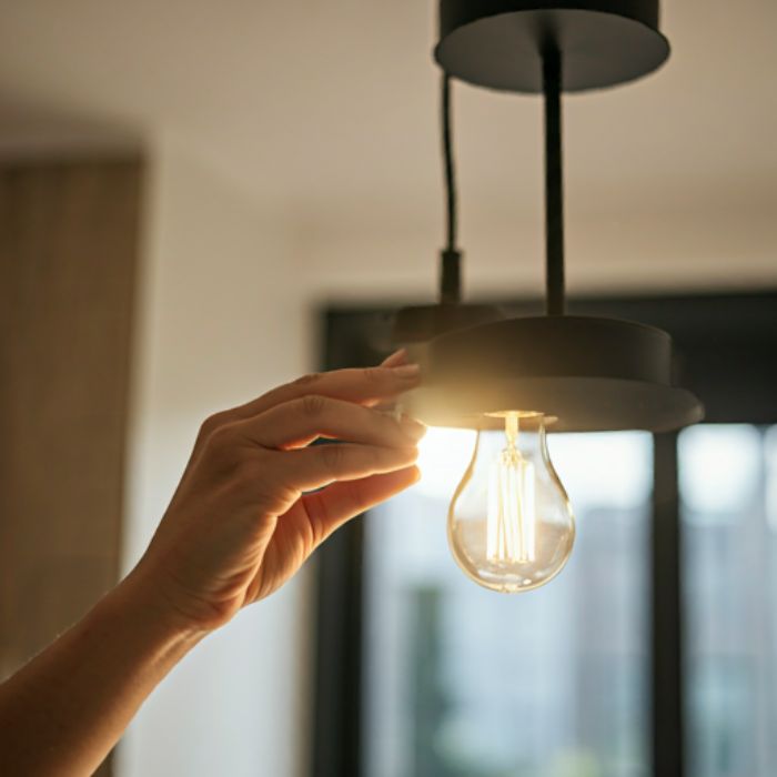 A person's hand changing a light bulb in a pendant light fixture. The light bulb is a warm-toned filament bulb.