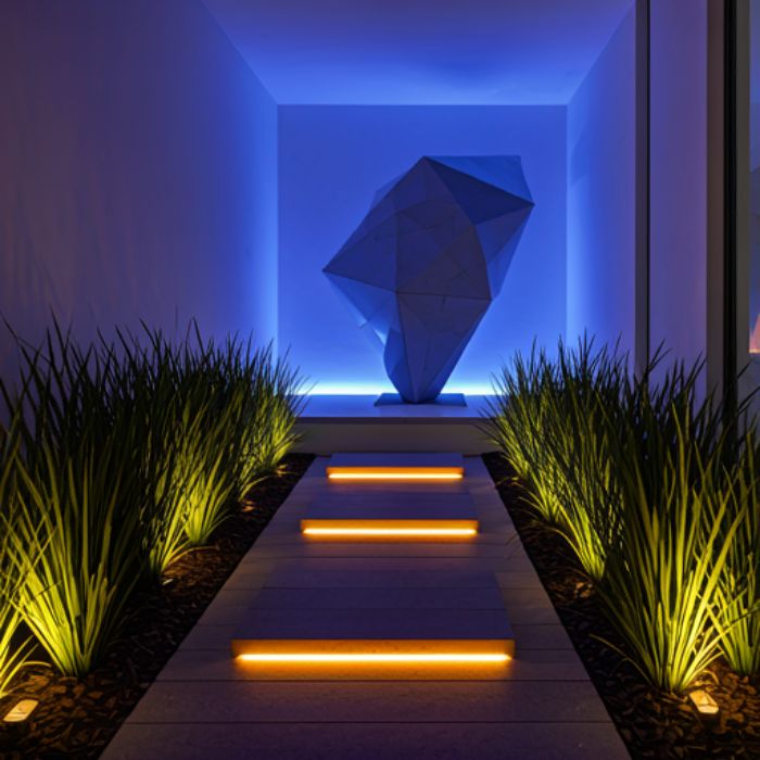 A modern entryway with a dramatic blue light illuminating a large, abstract sculpture. The pathway is lined with plants and illuminated by warm, orange lights.