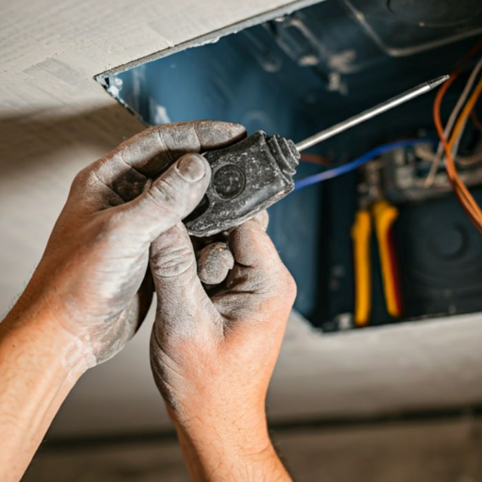 A close-up photo of an electrician's hands connecting wires to a junction box in a ceiling. The electrician's hands are covered in dust, and the image captures the detail of the tools and the electrical work being done.