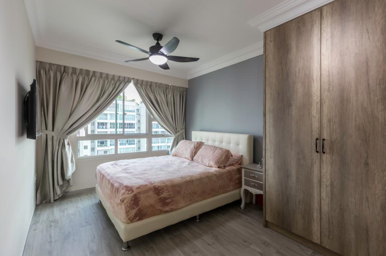 A cozy bedroom with a bed featuring a soft, pink comforter and a white headboard. The room has a dark gray accent wall, wooden flooring, and a ceiling fan with a light fixture.