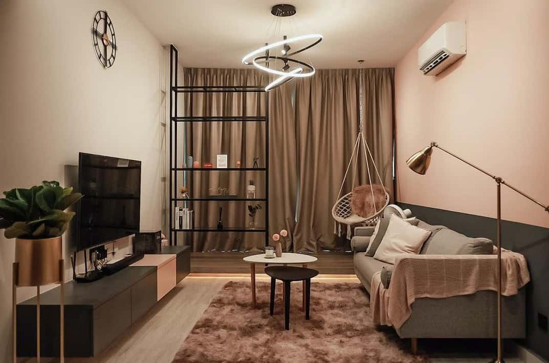 A cozy living room with a bohemian vibe. The room features a plush sofa, a hanging chair, a black metal shelving unit, and a unique circular pendant light. The color palette is warm and inviting with a touch of pink.