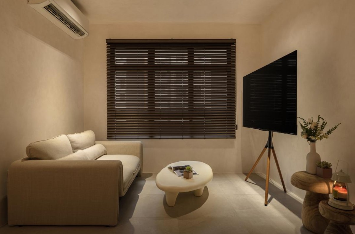 A cozy living room with a neutral color palette. The room features a comfortable sofa, a coffee table, a TV mounted on a tripod stand, and warm ambient lighting. The overall aesthetic is minimalist and inviting.