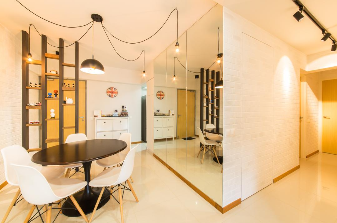 A modern dining room with a round wooden table and white chairs. The room features a unique divider with shelves and exposed Edison bulbs hanging from the ceiling, creating an industrial and stylish atmosphere.