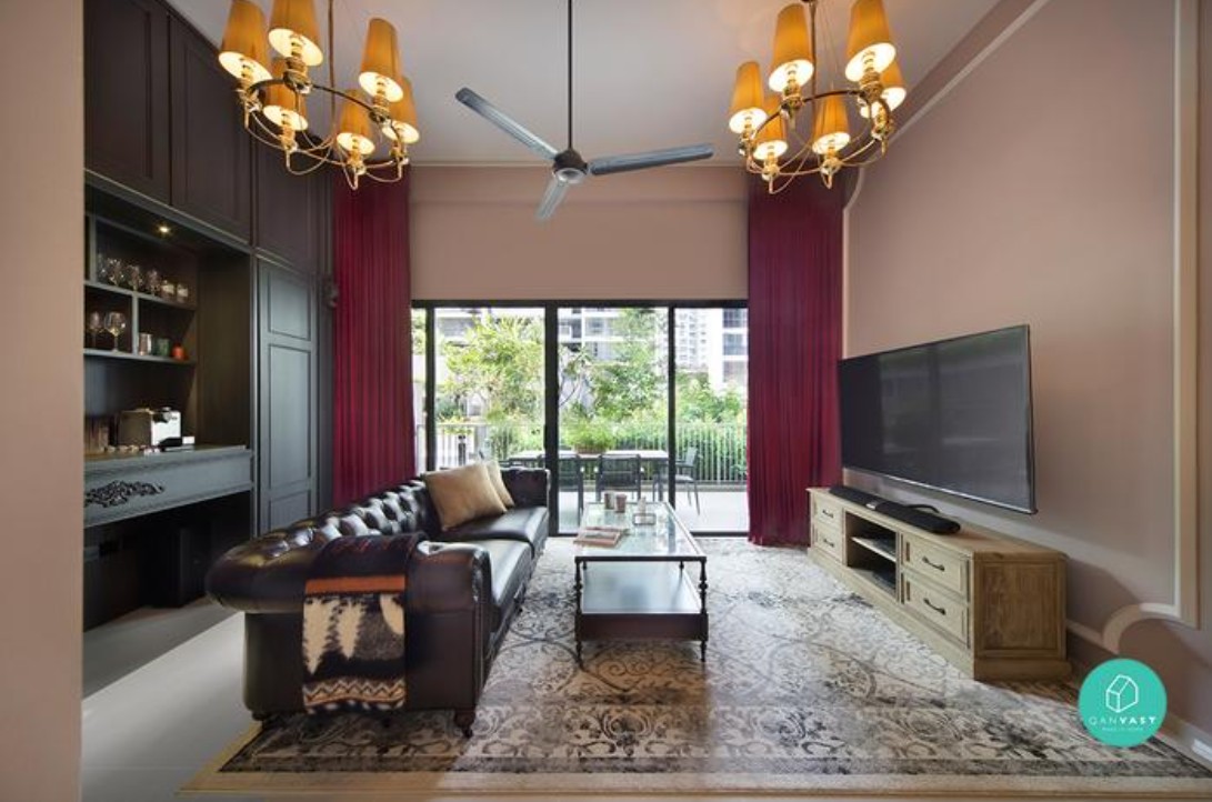 A luxurious living room with a leather sofa, a coffee table, and a large TV. Two vintage-style chandeliers with multiple light bulbs hang from the ceiling, adding a touch of elegance to the space. The room also features a patterned rug, dark red curtains, and a wooden TV stand.