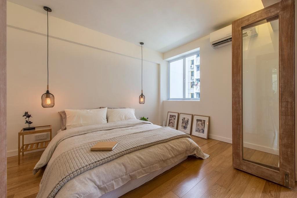 A minimalist bedroom with a neutral color palette. The room features a comfortable bed with white linens, wooden flooring, and pendant lights hanging over the bedside tables. The overall aesthetic is calm and inviting.