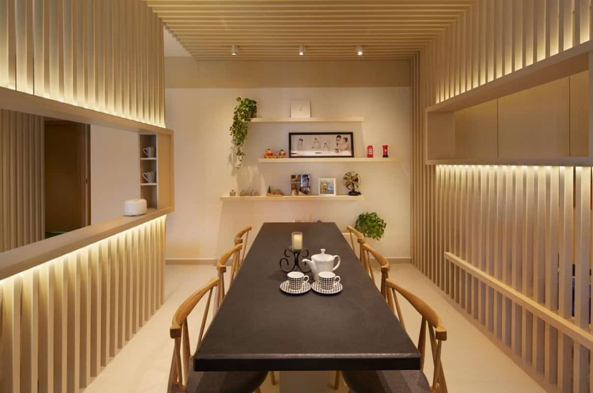 A minimalist dining room with a long, wooden dining table and chairs. The room features warm, wooden slatted walls and recessed lighting, creating a cozy and inviting atmosphere.