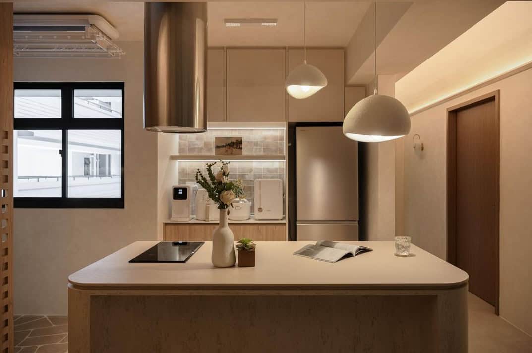 A minimalist kitchen with a warm, neutral color palette. The kitchen features a large island with a white countertop, wooden cabinetry, and pendant lights hanging above. The overall aesthetic is clean, functional, and inviting.