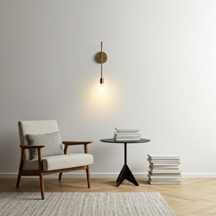 A modern living room with a sleek, brass wall sconce mounted on a white wall. The sconce features a minimalist design with a single light bulb, providing a warm and inviting glow. The room also includes a comfortable armchair, a side table, and a stack of books.