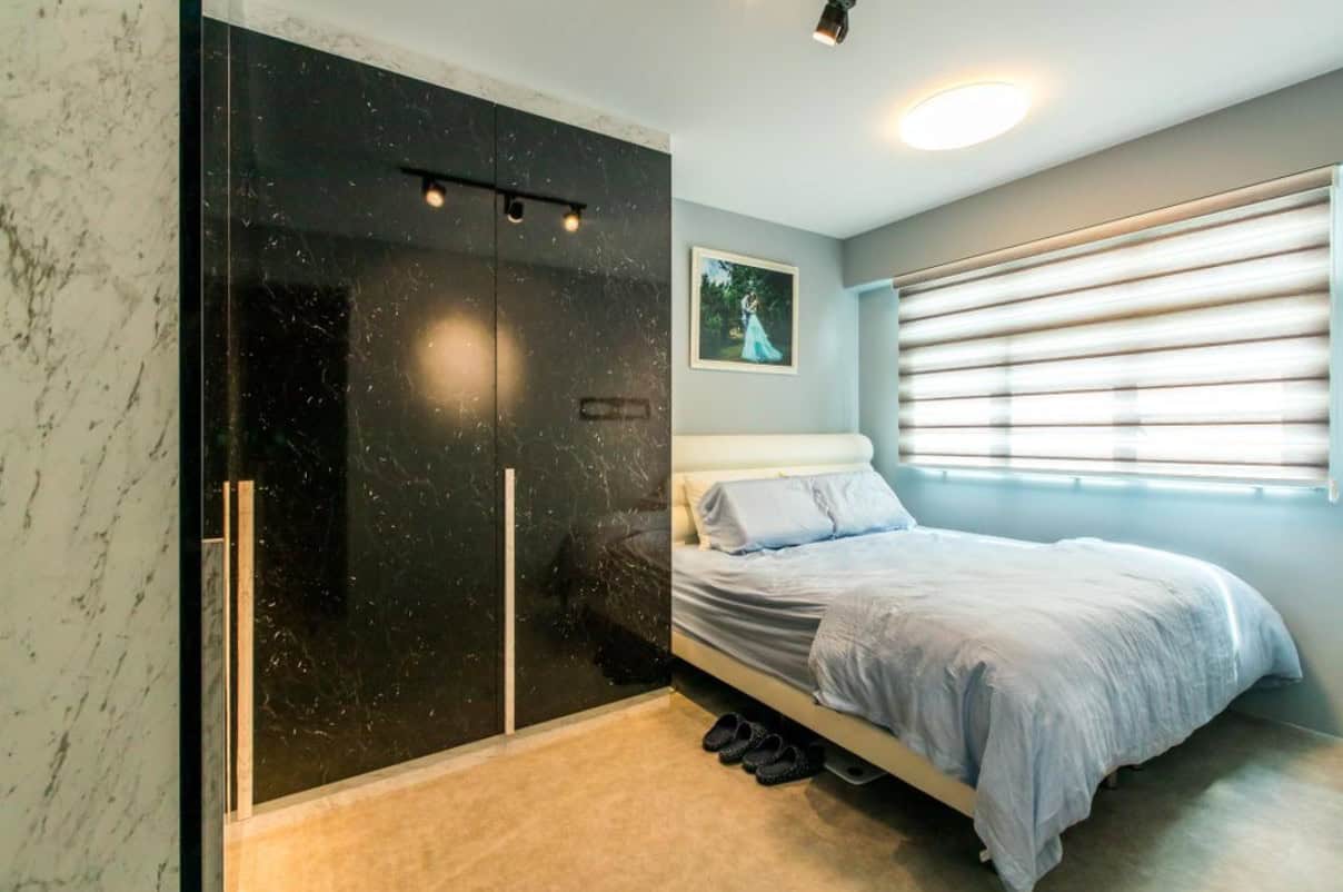 A modern bedroom with a minimalist design. The room features a large bed with a blue duvet, a black wardrobe with a mirrored door, and a large window with blinds. The overall aesthetic is clean, contemporary, and functional.