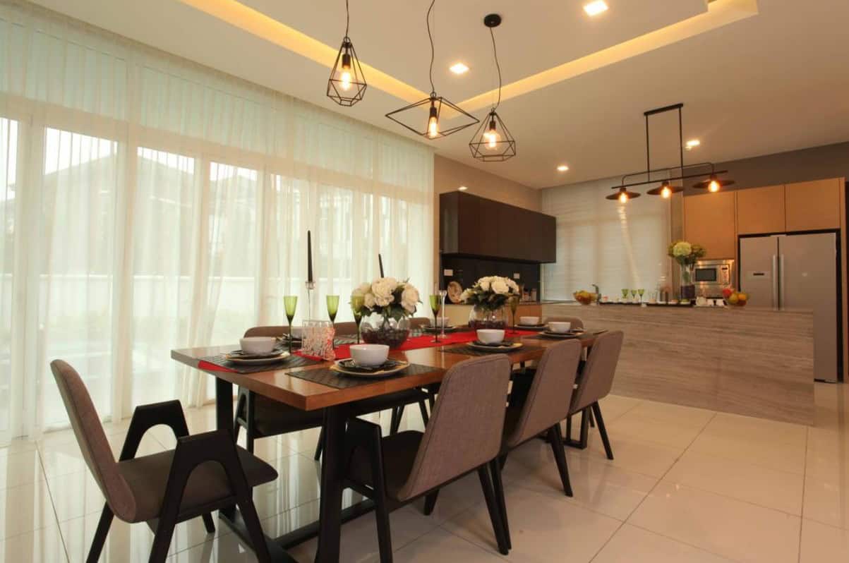 A modern dining area with a long wooden table and chairs. The room features large windows with sheer curtains, recessed lighting in the ceiling, and stylish pendant lights hanging above the table.