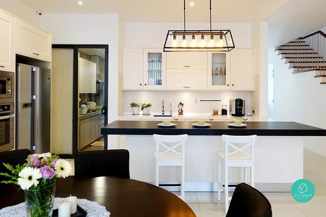 A modern kitchen with white cabinets and a black countertop. A breakfast bar with white stools sits adjacent to the kitchen, creating a casual dining area. A chandelier hangs above the breakfast bar, adding a touch of elegance.