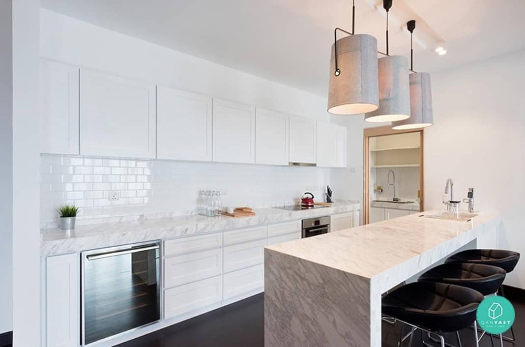 A modern kitchen with white cabinets, a marble countertop, and a breakfast bar with black bar stools. The kitchen features sleek pendant lights and a contemporary design aesthetic.