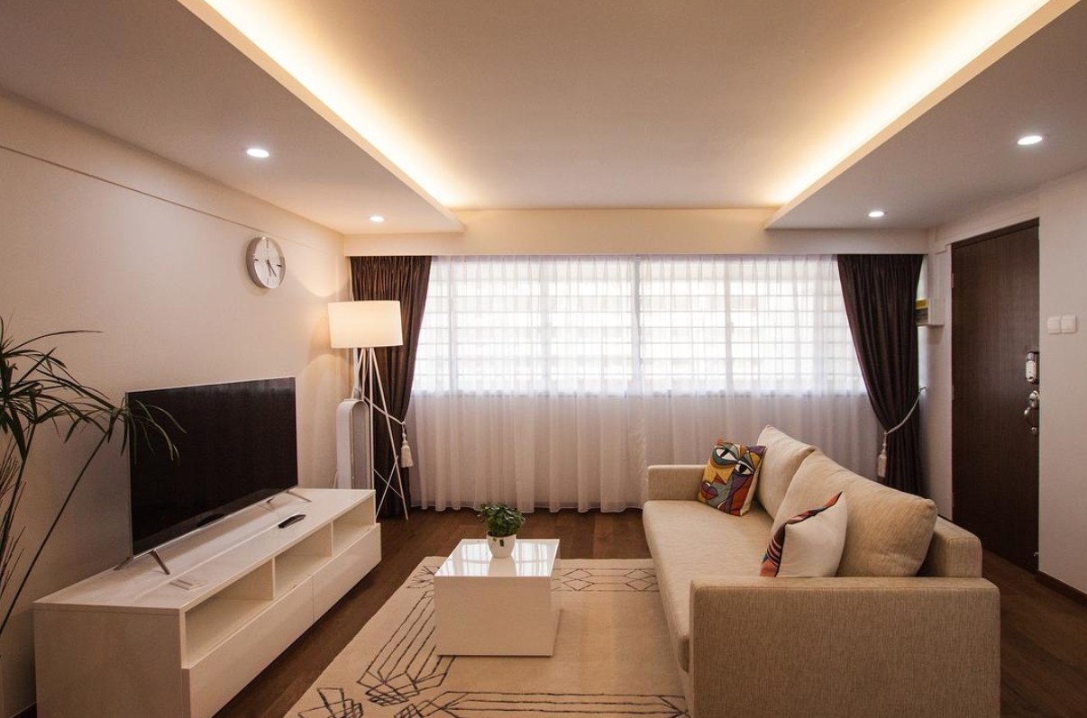 A modern living room with a cozy atmosphere. The room features recessed cove lighting, a comfortable sofa, a flat-screen TV, and large windows with sheer curtains.