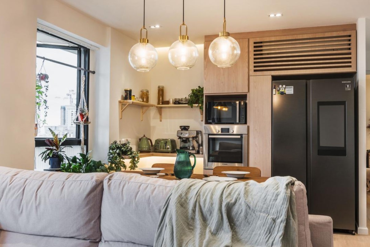 A modern, open-plan kitchen and living room with a minimalist design. The space features a light-colored sofa, a wooden dining table, and a sleek kitchen with stainless steel appliances. Three pendant lights with glass globes hang above the dining table, providing warm, ambient lighting.