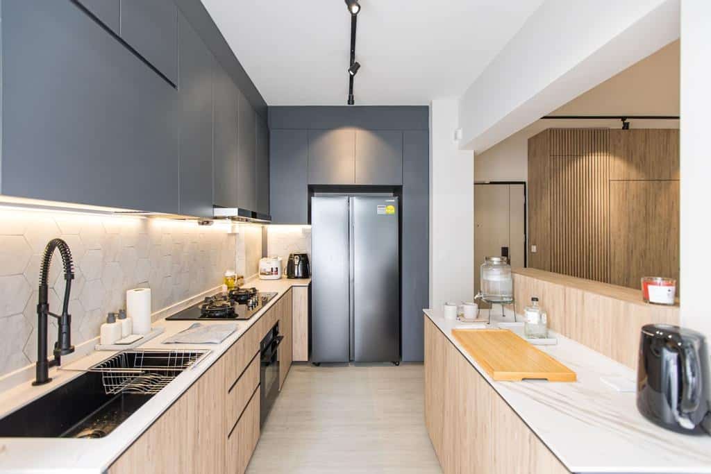 A modern, minimalist kitchen with a sleek design and warm wood accents. The kitchen features a long, L-shaped countertop with a black sink and faucet, a stainless steel refrigerator, and a white backsplash. The overall aesthetic is clean, functional, and inviting.