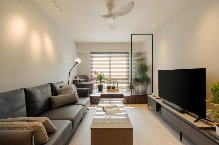 A modern, minimalist living room with a sleek gray sofa, a glass partition separating the living area from a smaller seating area, and a flat-screen TV mounted on the wall. The room is decorated with plants, and the overall aesthetic is clean and contemporary.