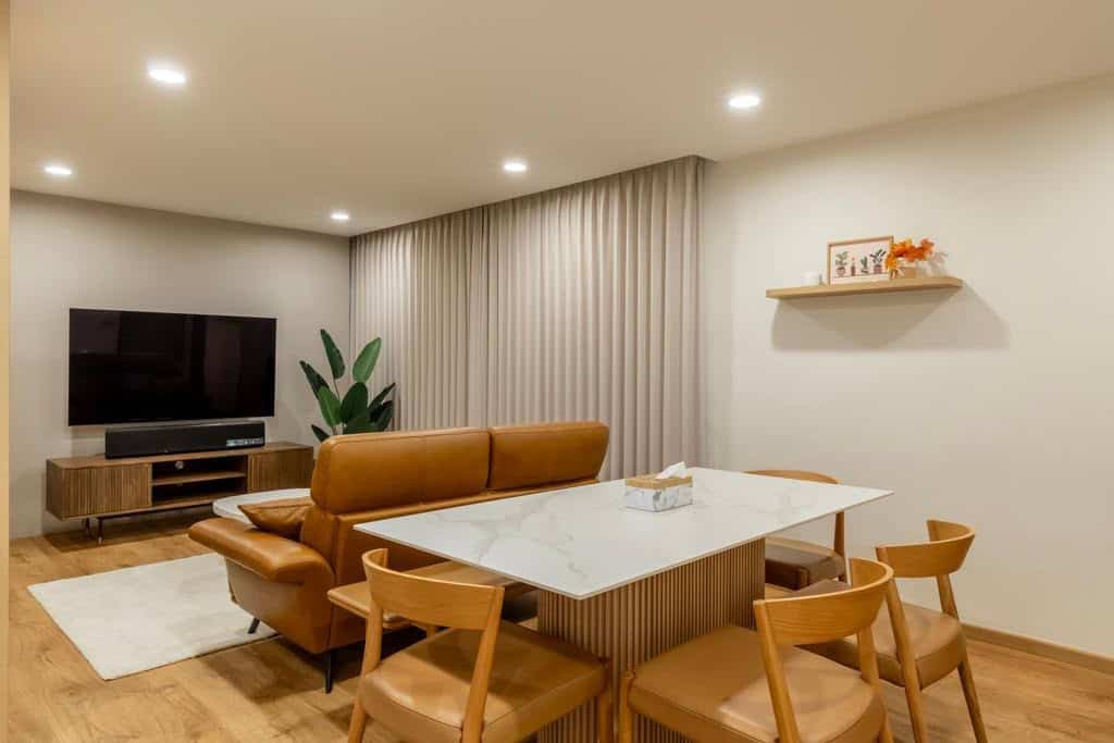 A modern open-concept living and dining area with a warm and inviting atmosphere. The space features a comfortable leather sofa, a dining table with wooden chairs, and a flat-screen TV. The overall design is minimalist and functional.