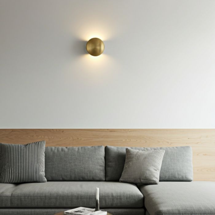 A modern living room with a sleek, brass wall sconce mounted on a white wall. The sconce features a minimalist design with a circular shape, providing a warm and inviting glow. The room also includes a gray sectional sofa, a coffee table, and a wooden accent wall.