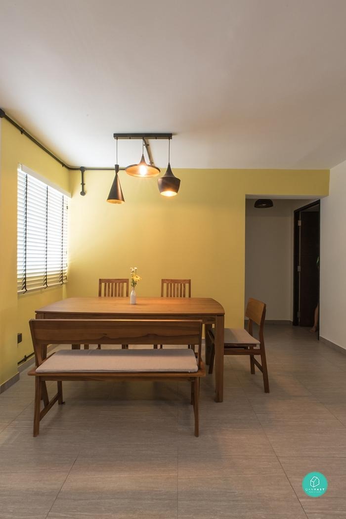 A dining room with a bright yellow wall and a wooden dining table. Three industrial-style pendant lights hang above the table, casting a warm glow over the space. The room has a cheerful and inviting atmosphere.